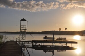 Un muelle de madera se extiende sobre aguas tranquilas, con una torre de salvavidas, bancos y globos aerostáticos a lo lejos.