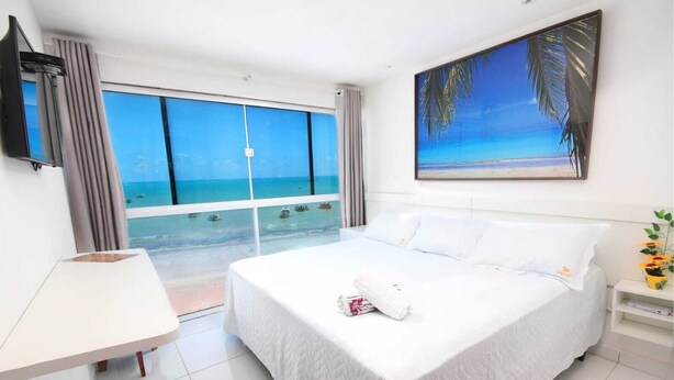 Une chambre d’hôtel avec un grand lit, un bureau et une vue sur l’océan grâce à de grandes fenêtres du sol au plafond.