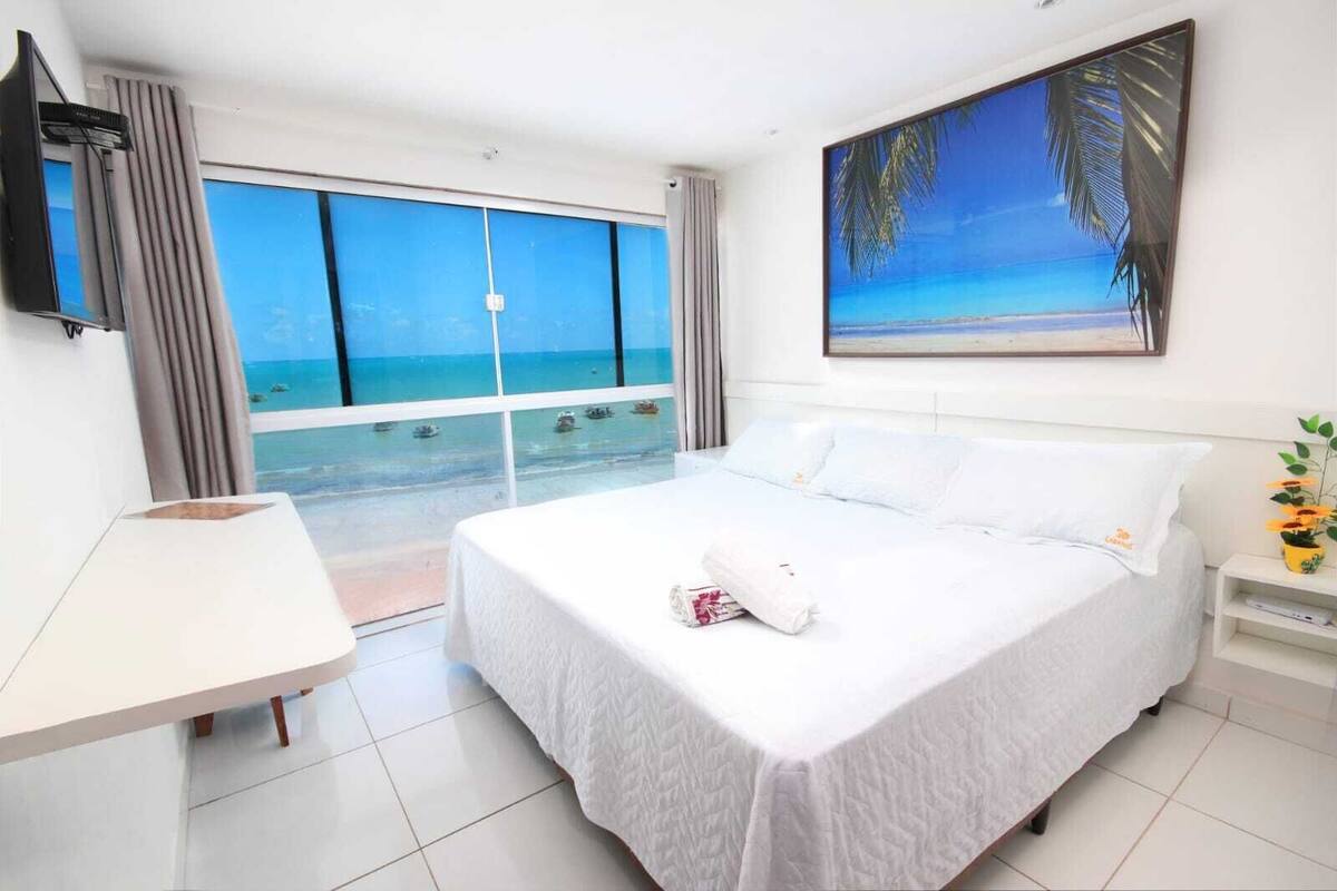 A hotel room with a large bed, a desk, and a view of the ocean through floor-to-ceiling windows.
