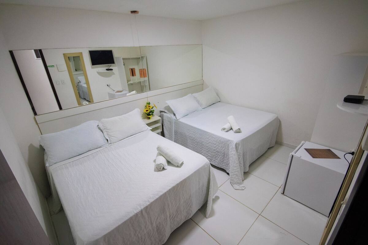A hotel room with two single beds, a large mirror, a small table with a vase of flowers, and a compact refrigerator.