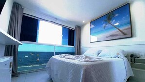 Une chambre à coucher avec un lit, un tableau représentant une plage, une fenêtre donnant sur la mer et un balcon.