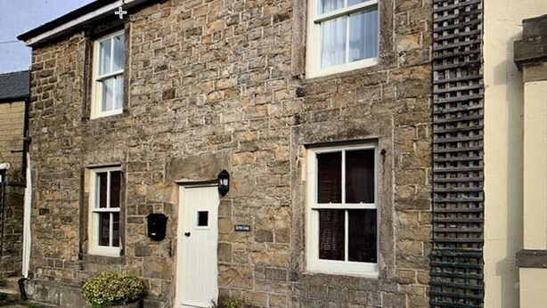 A stone building with a white door, two windows with shutters, and a small garden area with flowers.