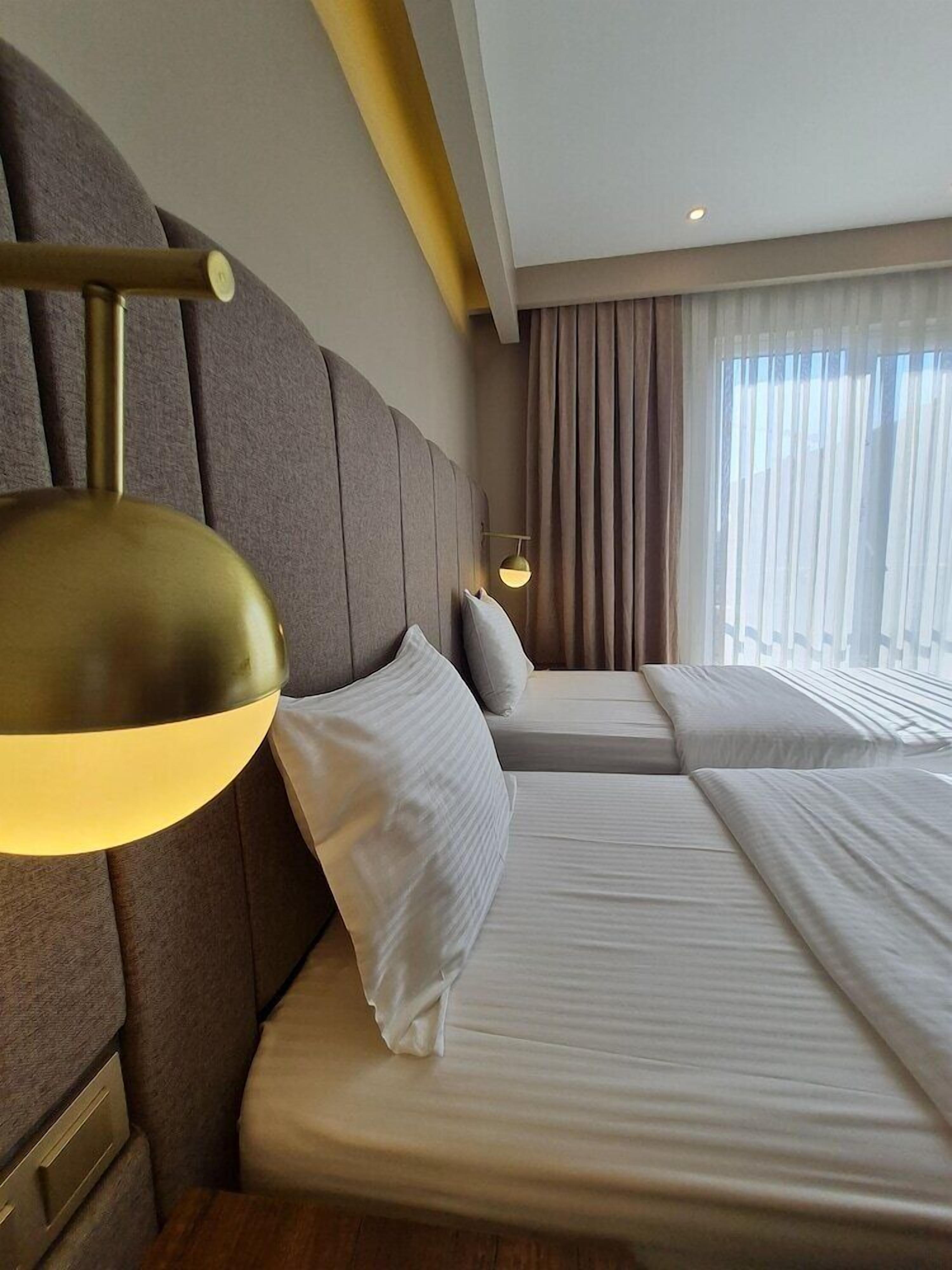 A hotel room with two beds, a headboard, a bedside lamp, a window with curtains, and a wall-mounted light fixture.