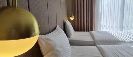 A hotel room with two beds, a headboard, a bedside lamp, a window with curtains, and a wall-mounted light fixture.