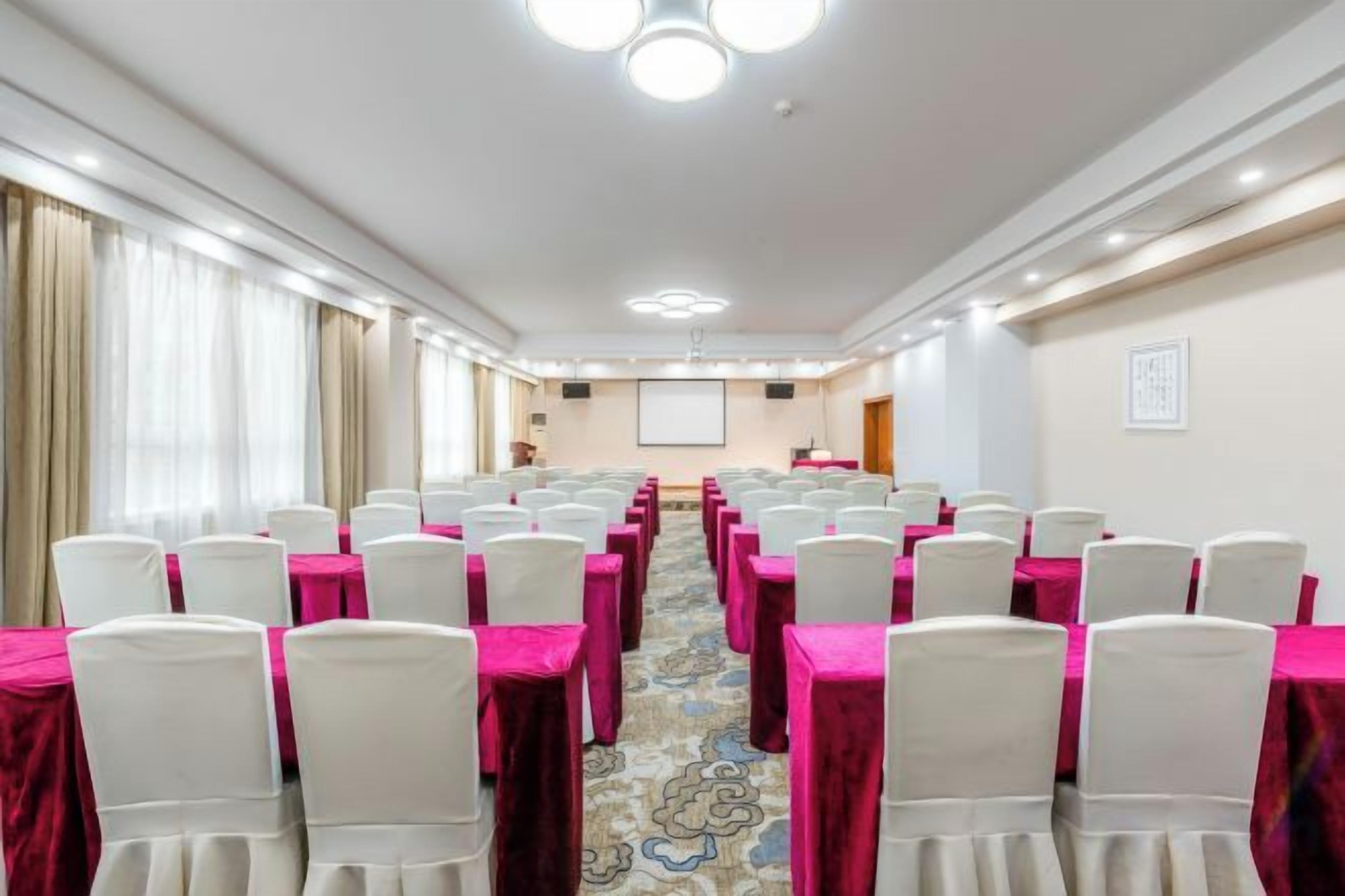 Sala de conferências com fileiras de cadeiras e mesas, uma tela de projetor e uma área para apresentações.