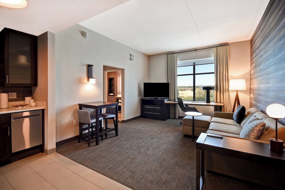 A modern hotel room with a kitchenette, a sofa, a TV, and a large window with curtains.