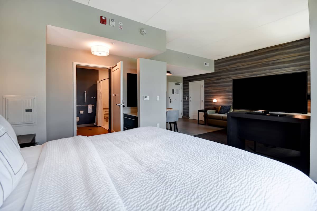 A hotel room with a large bed, a TV on a stand, a desk, and a bathroom visible through an open door.