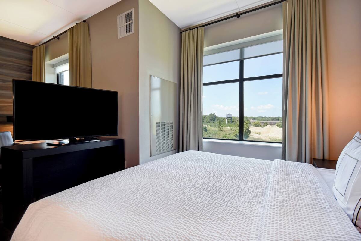 A hotel room with a large bed, a flat-screen TV on a dark dresser, and a window with curtains.