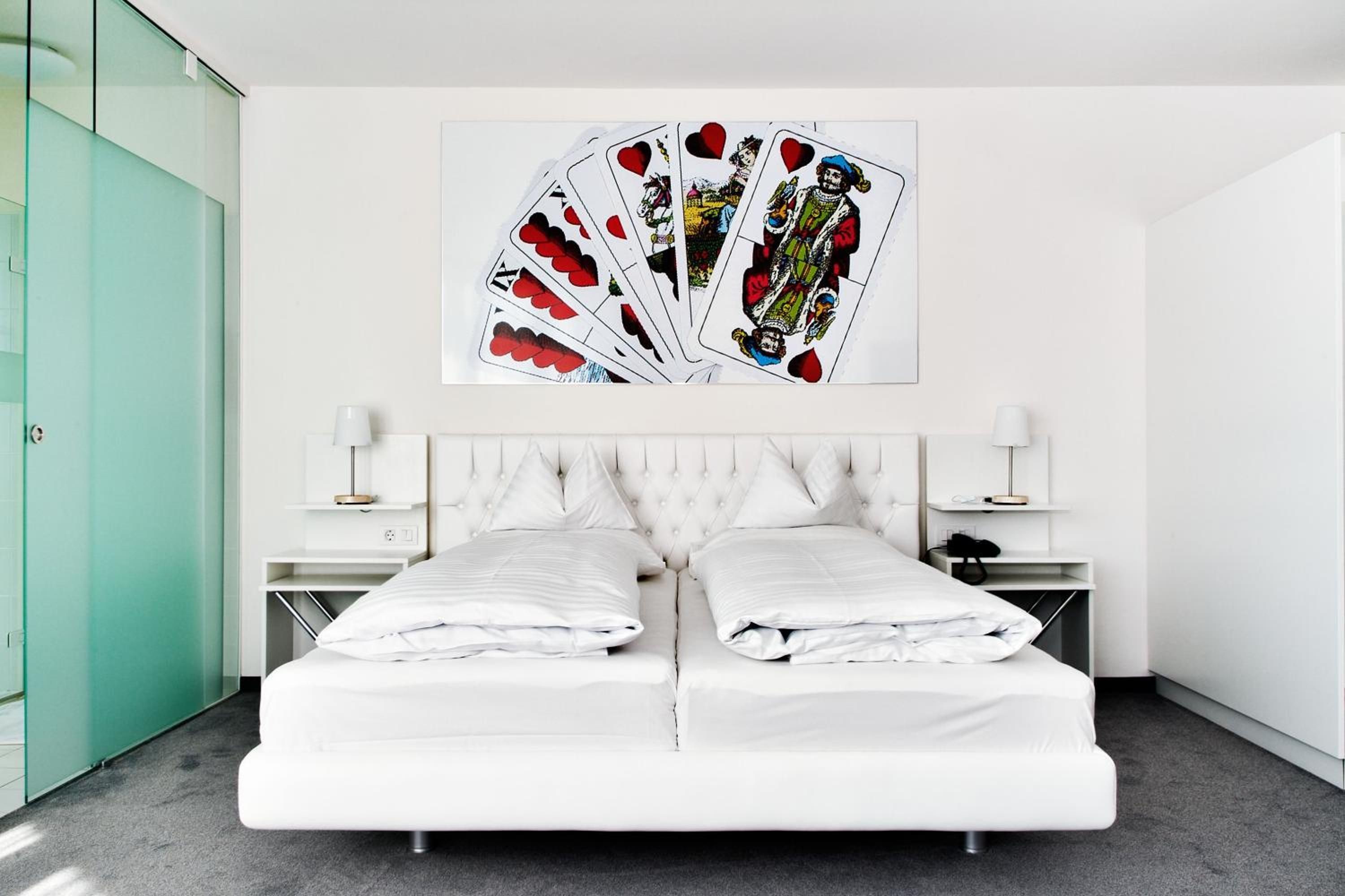 Habitación de hotel con una cama, dos mesitas de noche, una obra de arte en la pared con cartas de juego y una puerta de vidrio verde.