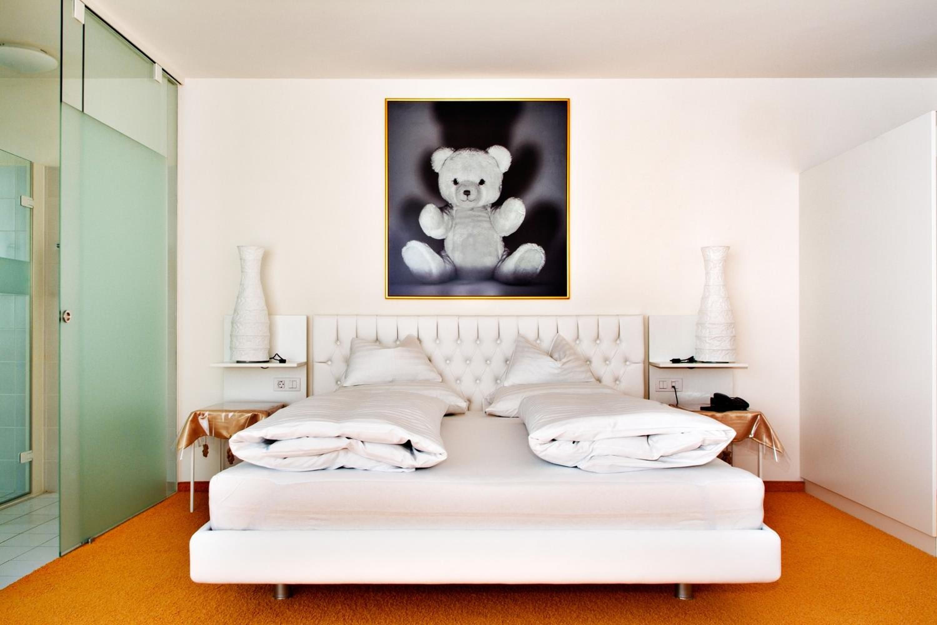Un dormitorio moderno con una cama grande, un cabecero acolchado, dos mesitas de noche con lámparas y un cuadro en la pared con un osito de peluche.