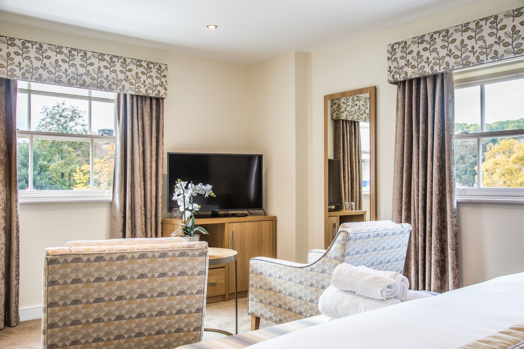 Een hotelkamer met een bed, twee fauteuils, een televisie, een spiegel en een raam met gordijnen.
