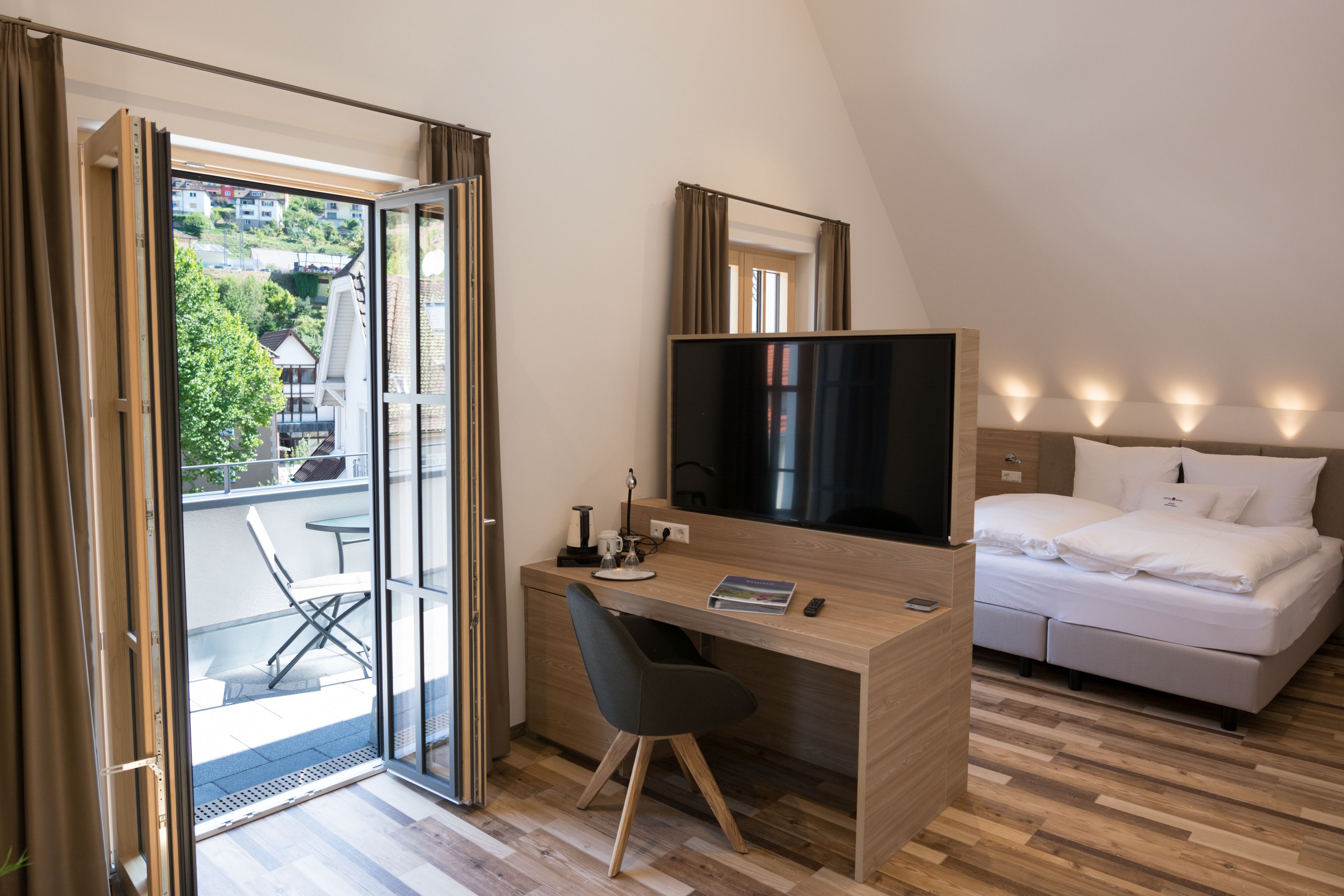 A hotel room with a wooden desk, a bed, a TV, and a balcony with a chair.