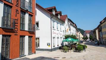 Un hotel con fachada moderna, un patio con mesas al aire libre y arquitectura europea tradicional en el fondo.