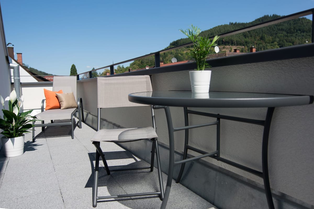 Una terraza en la azotea con una mesa y sillas, plantas en macetas y vistas del paisaje circundante.