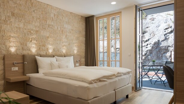 A modern bedroom with a large bed, a wooden headboard, a nightstand, and a view of a balcony with outdoor seating. - Hotelmyhome (Hornberg)