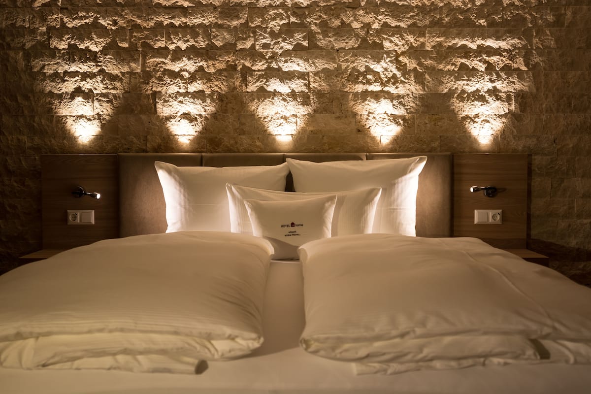 Una cama bien tendida con sábanas y almohadas blancas, un cabecero de madera y una pared de piedra texturizada al fondo.
