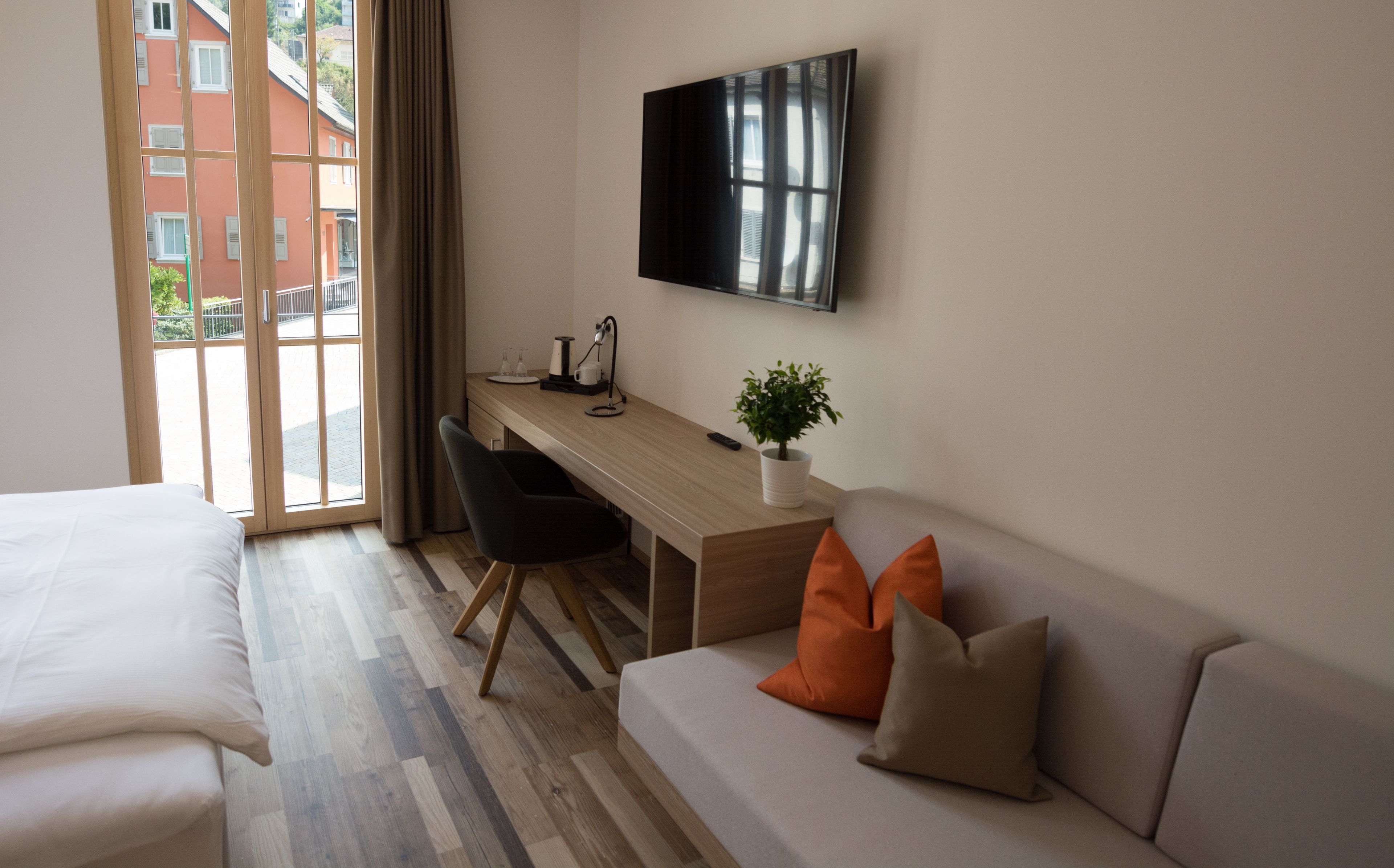 A hotel room with a bed, a sofa, a desk with a plant, a TV, and a view of buildings through the window.