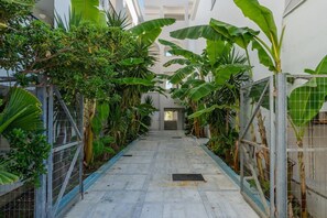 A corridor with a tiled floor, flanked by metal fences and lush green plants, leading to a door at the end.
