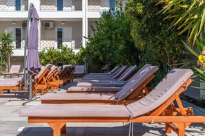 A row of sun loungers with a covered umbrella, arranged in front of a building with balconies.