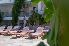 A poolside area with lounge chairs and umbrellas, surrounded by greenery and a building with balconies.