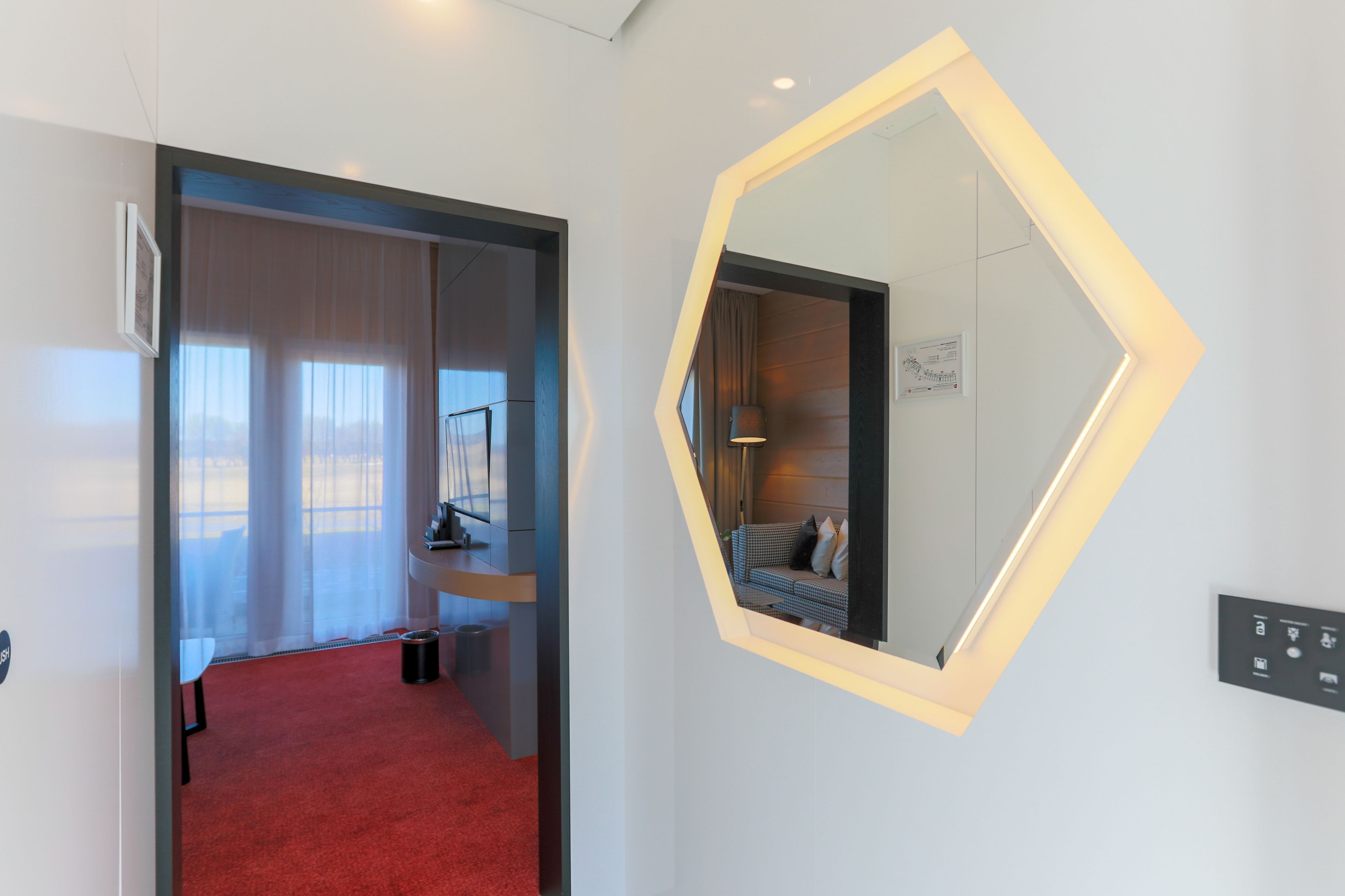 Une chambre d’hôtel avec un tapis rouge, un grand miroir doté d’un cadre géométrique et une vue sur un balcon agrémenté de rideaux légers.