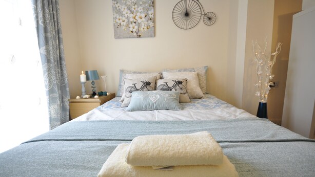 Een net opgemaakt bed met een fietsen-thema kussen, een aan de muur bevestigde fietsdecoratie en een vaas met takken.