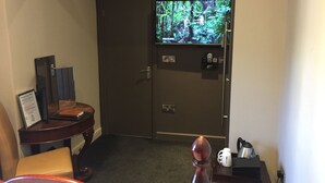 A hotel room with a dark wooden door, a television displaying a nature scene, a small wooden side table with a framed picture, a beige chair, and a coffee maker on a tray. - The Peacock Inn (Bakewell)
