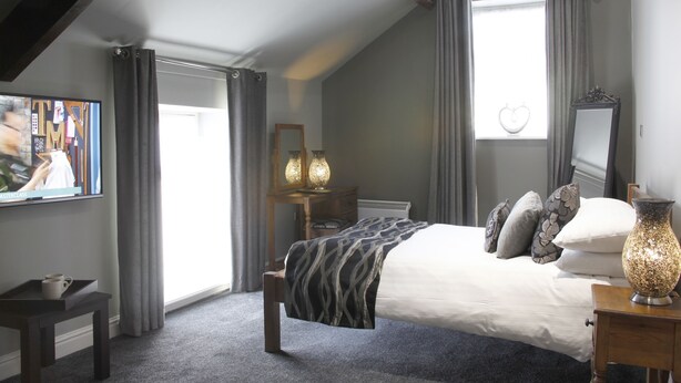 A bedroom with a large bed, a bedside table, a mirror, and a window with curtains. - The Peacock Inn (Bakewell)