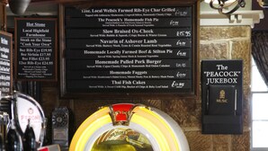 A bar area with a jukebox, a menu board listing food and drink items, and a framed sign that reads "The Peacock Jukebox." - The Peacock Inn (Bakewell)