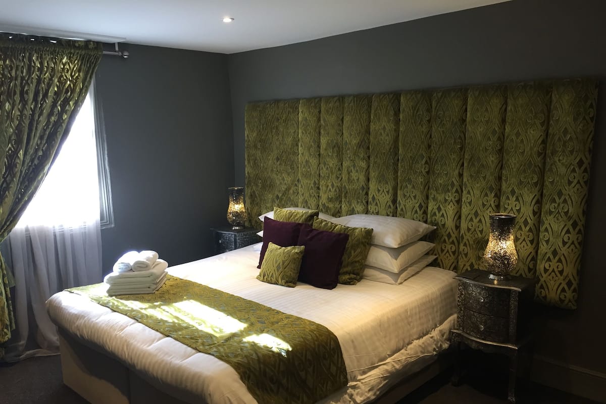 A neatly made bed with a green throw, pillows, and a folded towel on a headboard. There are two bedside tables with lamps.
