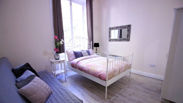 A bedroom with a bed, a bedside table with a lamp, a dresser, and a window with curtains. - Paddington Guest Apartment (London)