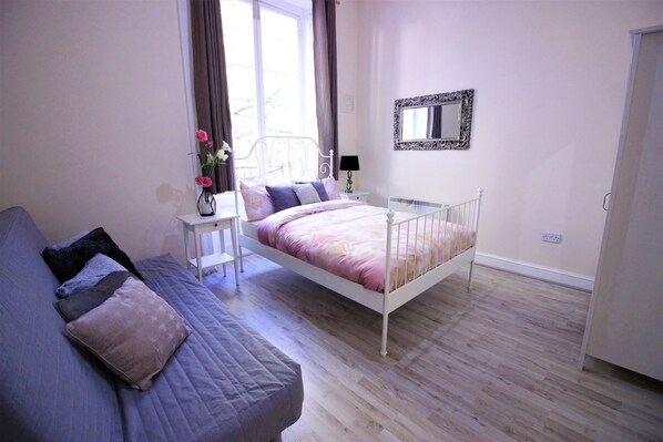 A bedroom with a bed, a bedside table with a lamp, a dresser, and a window with curtains. - Paddington Guest Apartment (London)