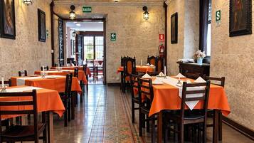 En restaurant med en lang, flisebelagt gulv, der fører hen til en dør mærket 'SALIDA'. Bordene er dækket med orange dugge, og væggene er prydet med indrammede billeder.