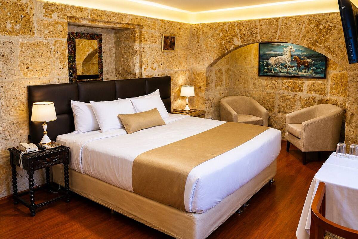 A hotel room with a large bed, a nightstand, a chair, a table, and a painting on the wall.