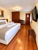 A hotel room with a bed, a desk, a chair, a television, and a wooden floor.
