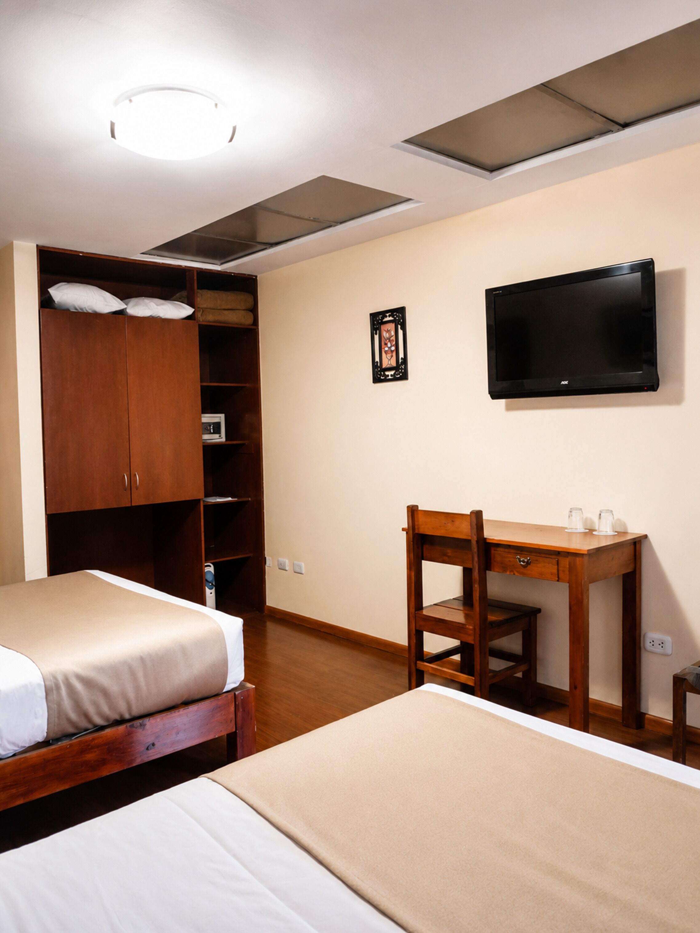 A hotel room with two beds, a desk, a television, a wardrobe, and a ceiling light.