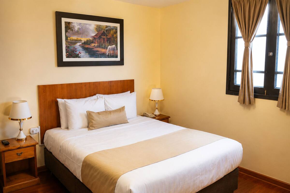 A hotel room with a large bed, wooden headboard, two bedside tables with lamps, a painting on the wall, and a window with curtains.