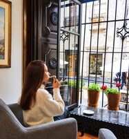 Une personne est assise près d’une fenêtre, avec des plantes en pot, un tableau encadré accroché au mur, et une vue sur la rue.