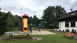 Een speeltuin met een gele glijbaan, banken en een gebouw op de achtergrond.