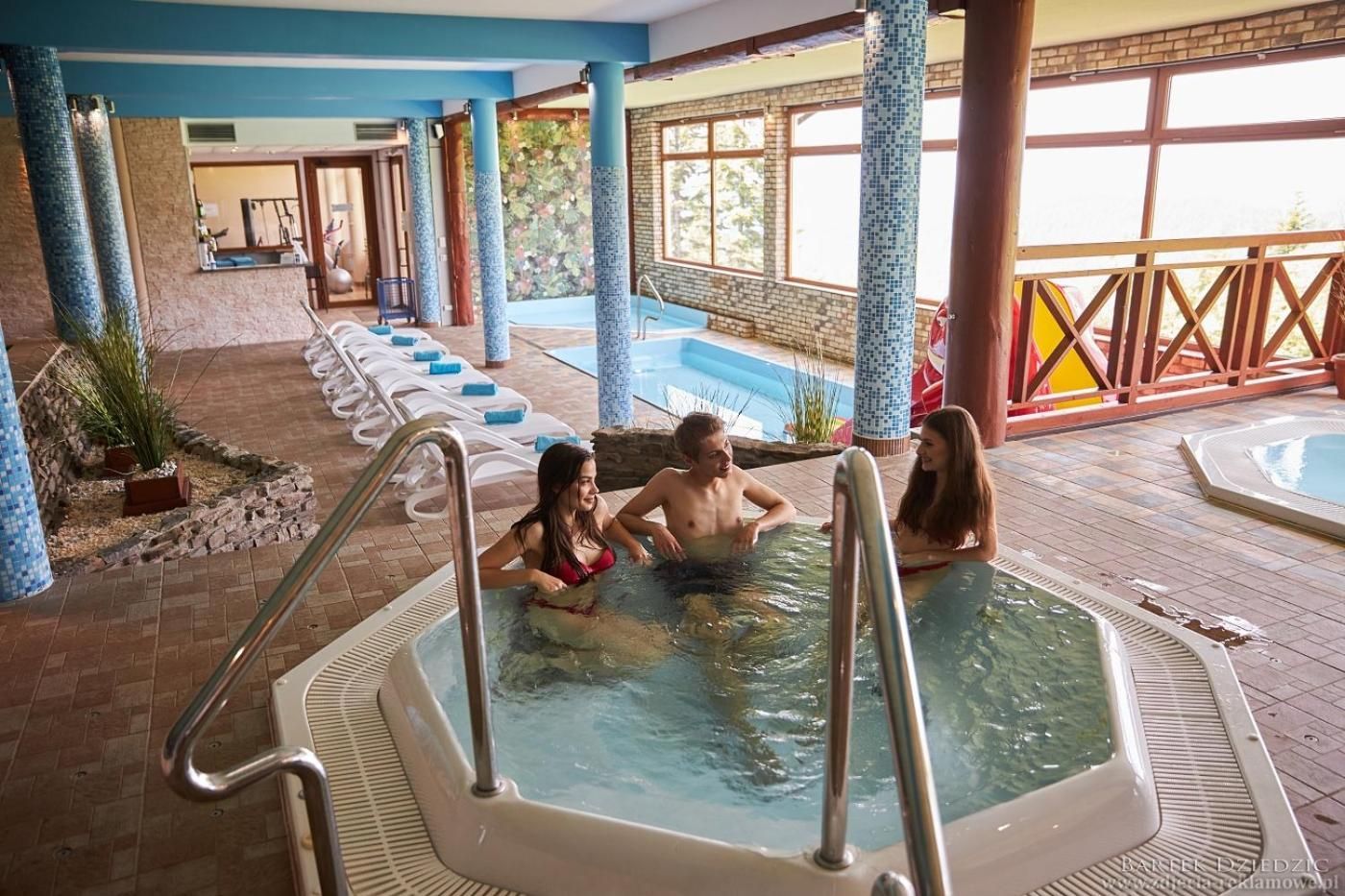 Un área de piscina interior con varias piscinas, sillas de descanso y un suelo de baldosas de piedra.