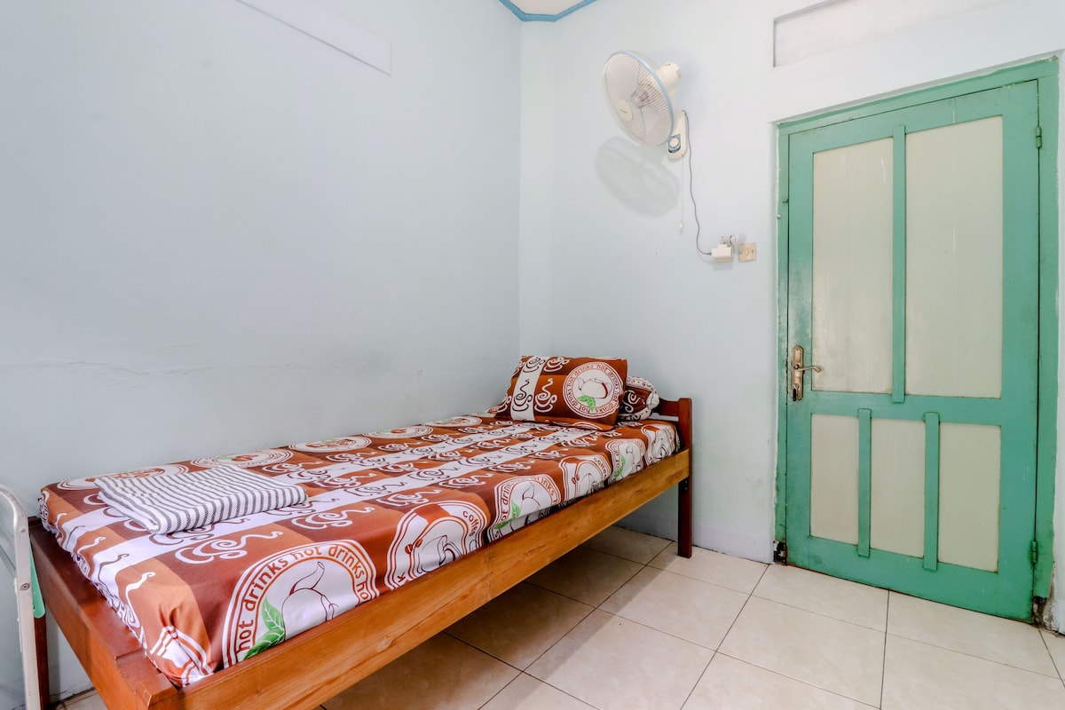 Ein Einzelbett mit gemusterten Betttextilien, ein Standventilator und eine grüne Tür in einem Zimmer mit weißen Wänden und Fliesenboden.