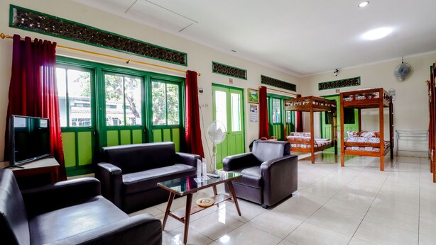 A hotel room with green-framed windows, a flat-screen TV, leather sofas, a wooden bunk bed, and a fan. - Bahagia Sederhana Home Stay - Hostel (Bantul)