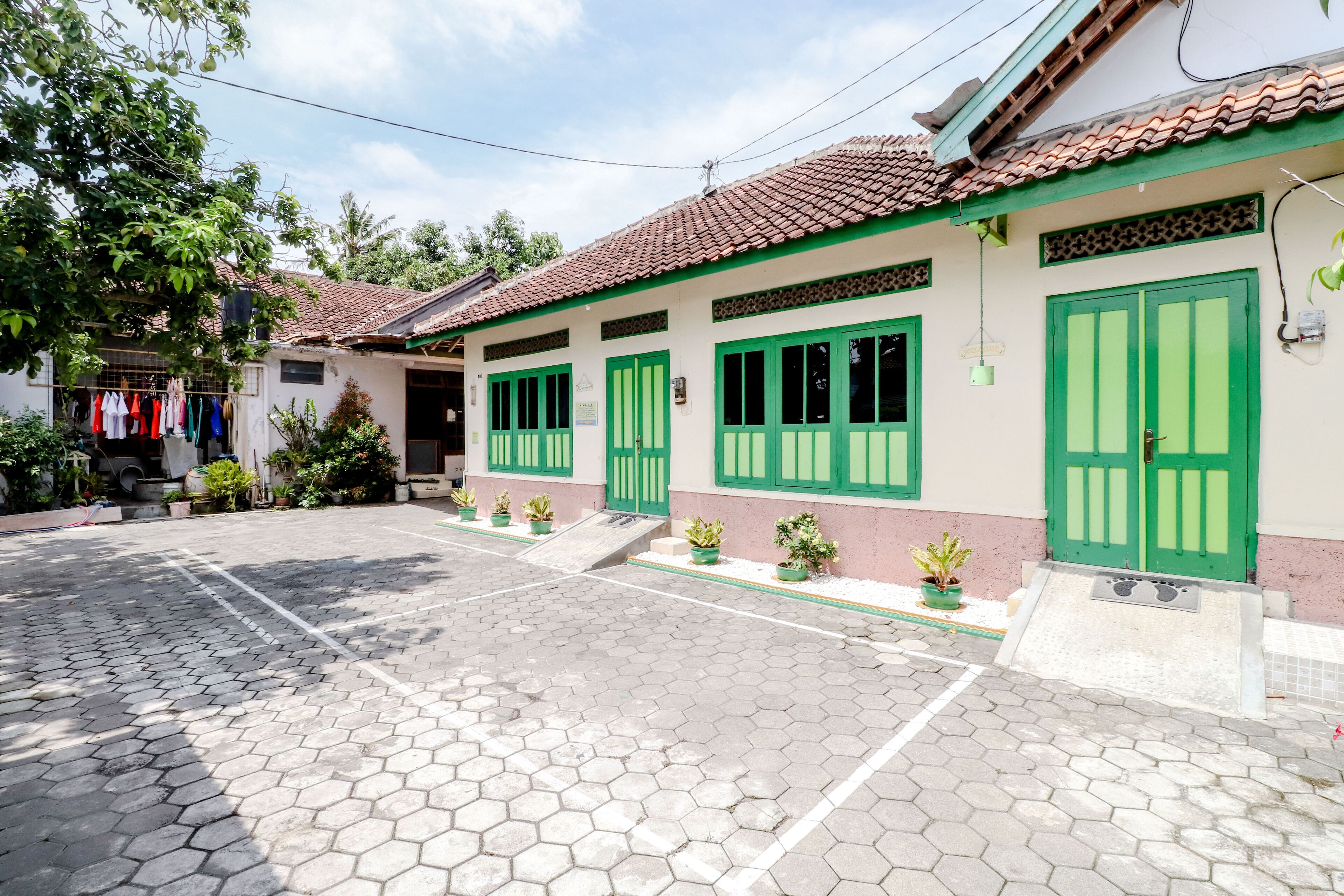 A building with green doors and windows, a tiled roof, and a paved courtyard with potted plants.