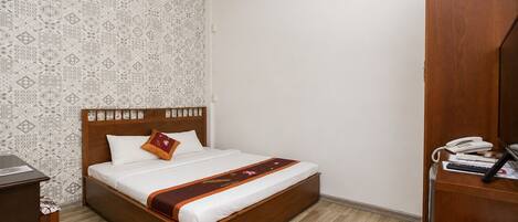 A hotel room with a wooden bed, a patterned wall, a wooden desk with a telephone, and a small table with a teapot.