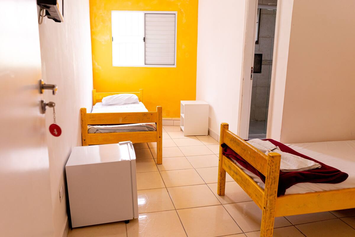 A room with two wooden beds, a white refrigerator, a white bedside table, and a window with blinds.