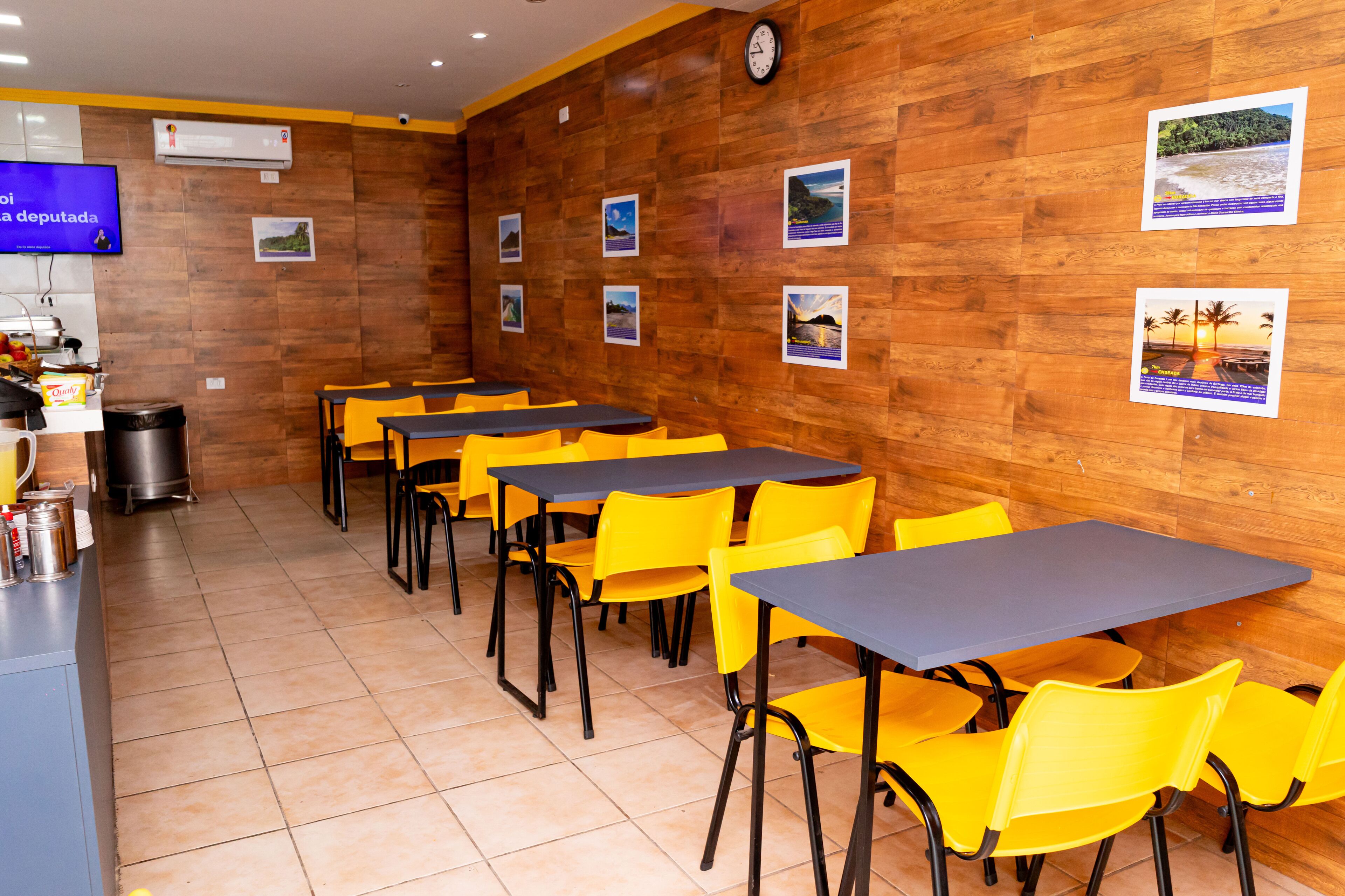 Um restaurante com paredes de madeira, cadeiras amarelas e mesas. Há quadros nas paredes e um relógio.