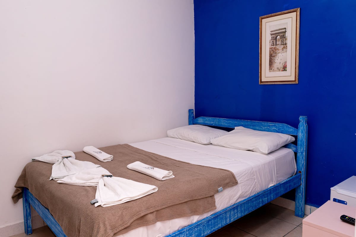 A single bed with a blue headboard, white bedding, and a brown blanket. There are towels on the bed and a framed picture on the wall.