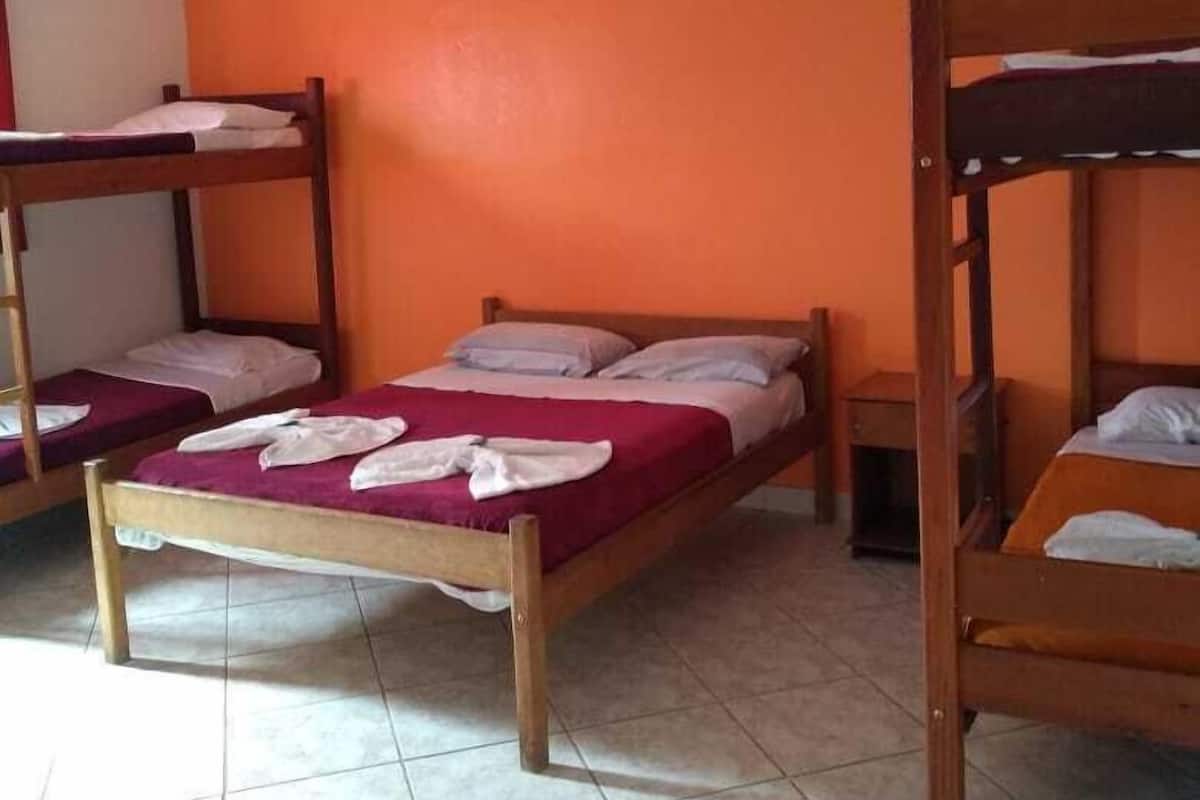 A dormitory room with three wooden bunk beds, each with a mattress and pillows. The room has orange walls and white tiled flooring.