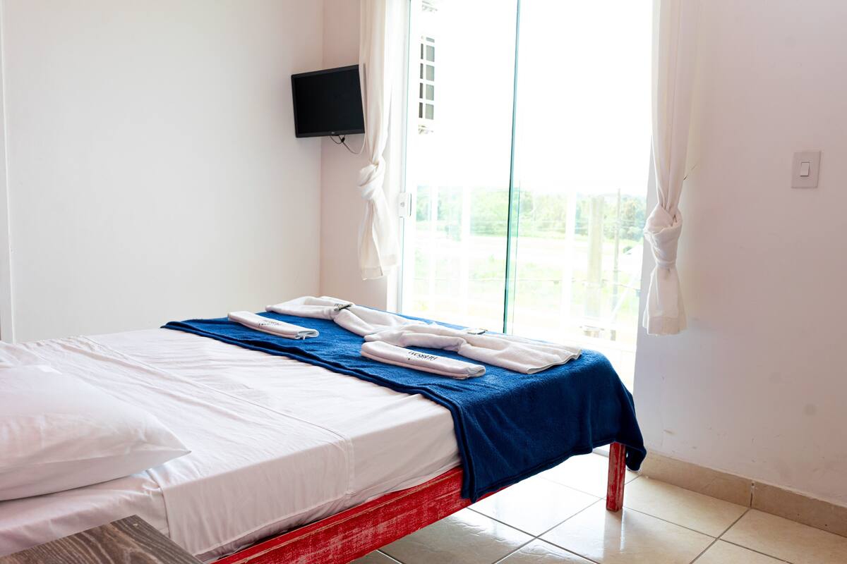 A hotel room with a bed, a TV mounted on the wall, and a view of the outdoors through a large window.