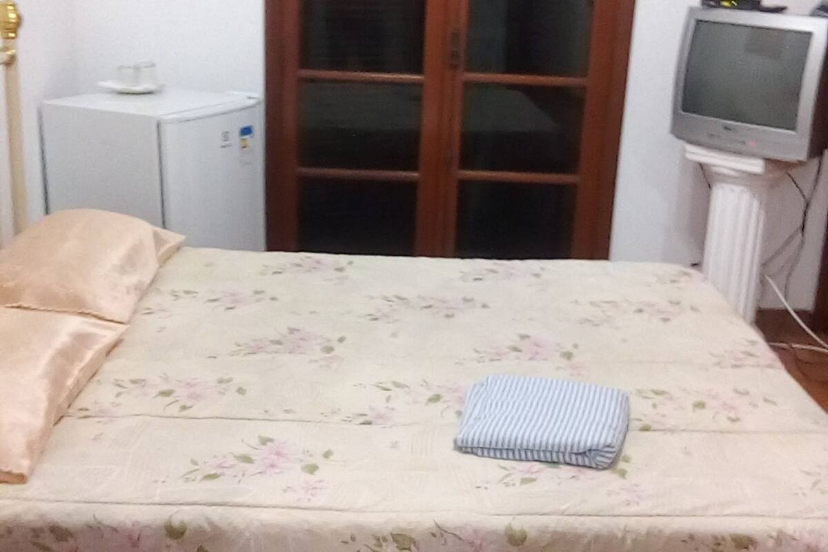 A bedroom with a bed, a wardrobe, a small white cabinet, and a television.